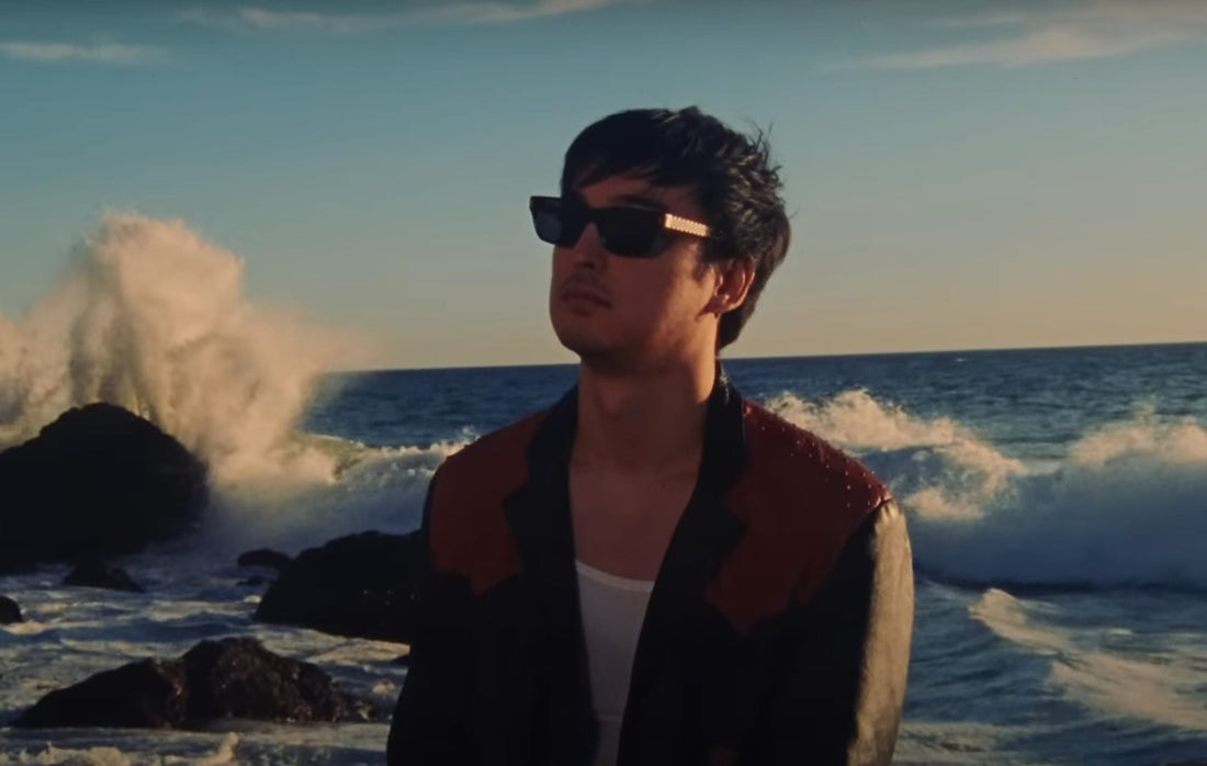 Joji standing by the ocean with waves splashing on boulders in the background.